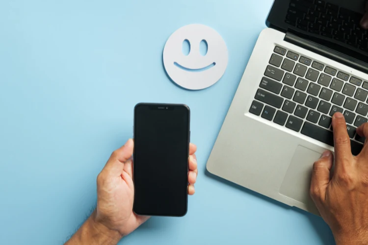 A customer's hands holding a smartphone next to a laptop and a smiley-face icon — a branded-search flow turns a QR scan into a legitimate engagement signal Google recognizes